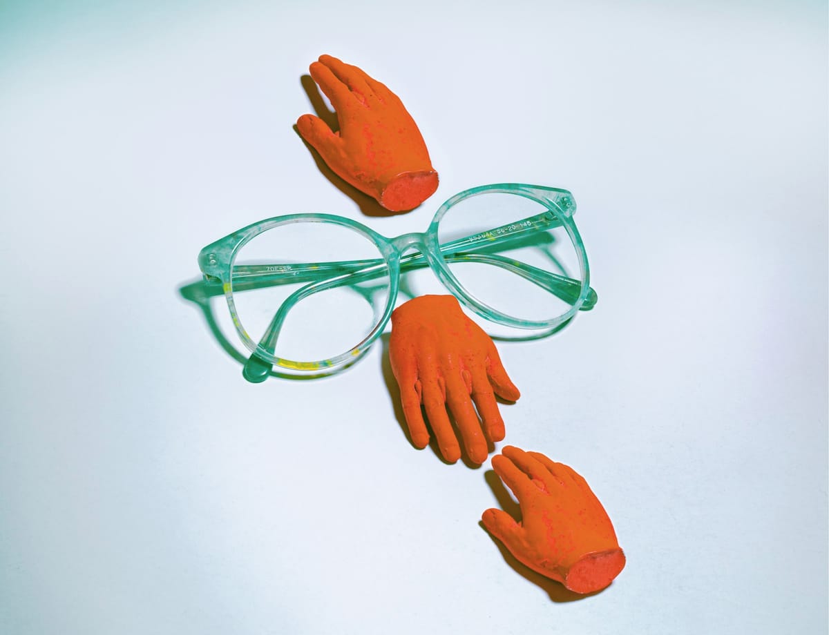 A pair a teal glasses on a white surface.  And Orange hand reaches up from the bridge, another down.  A third hand of the same color reaches toward the second