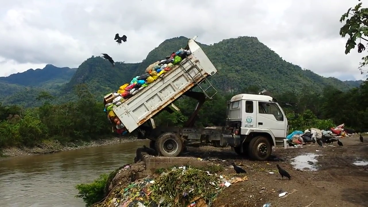 A photo of a dump truck pouring trash into a river while carrion birds circle it.