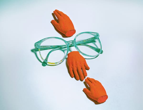 A pair a teal glasses on a white surface.  And Orange hand reaches up from the bridge, another down.  A third hand of the same color reaches toward the second